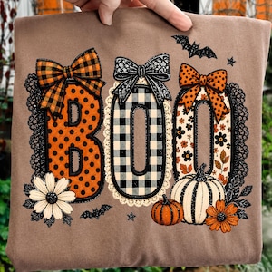 May include: A brown t-shirt featuring the word BOO in large letters. The B has orange polka dots and a plaid bow, the O has a black and white buffalo check pattern and a black lace bow, and the second O has a floral print and an orange polka dot bow.