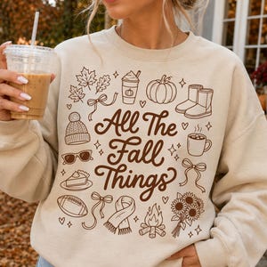 May include: A beige crewneck sweatshirt featuring brown line art illustrations of autumn-themed items including a pumpkin, boots, a scarf, a campfire, sunflowers, and a football, with the text All The Fall Things printed in the center.