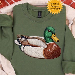 Peut inclure: Un sweat-shirt vert olive avec une broderie de canard colvert. Le canard a une tête verte, une poitrine marron et des ailes blanches et marron. Une étoile jaune dans le coin supérieur droit indique "9 Bonus Designs Included".