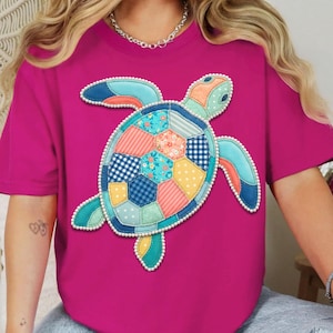 May include: A magenta t-shirt featuring a colorful patchwork turtle design. The turtle has a variety of patterned sections in blue, orange, yellow, and green, outlined with white beads. The shirt has short sleeves and a crew neck.