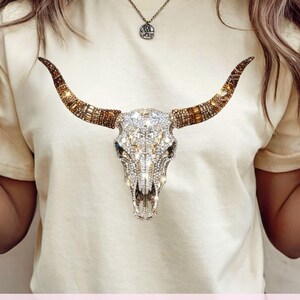 May include: A cream-colored t-shirt featuring a central graphic of a bull skull adorned with sparkling clear and amber-toned rhinestones. The skull design includes long, curved horns decorated with brown and gold crystals. Text reads: DIGITAL DOWNLOAD - NOT A PHYSICAL ITEM.