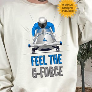 Puede incluir: Sudadera de color crema con una ilustración de dibujos animados de un bobsleigh y un piloto en azul y plata. El texto "FEEL THE G-FORCE" está impreso en azul y gris. Un gráfico de ráfaga amarilla dice "9 Bonus Designs Included."