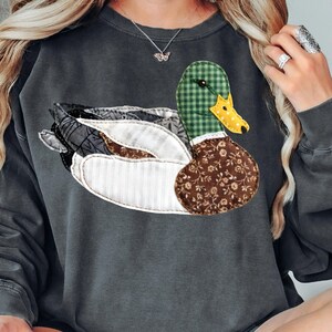 May include: Dark gray sweatshirt featuring a large, appliqued duck design. The duck has a green plaid head, a yellow beak, and a body made of various patterned fabrics in shades of brown, white, and gray. The sweatshirt is a casual style.