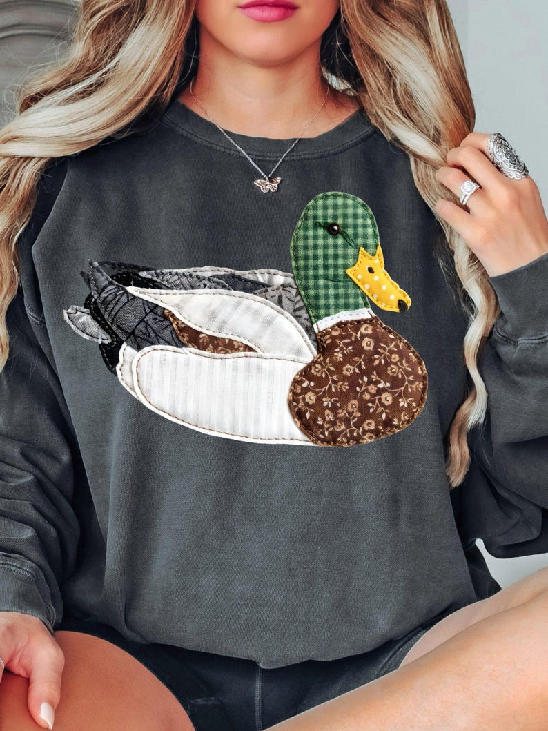 May include: Dark gray sweatshirt featuring a large, appliqued duck design. The duck has a green plaid head, a yellow beak, and a body made of various patterned fabrics in shades of brown, white, and gray. The sweatshirt is a casual style.