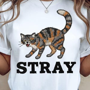 以下が含まれることがあります： 白地のTシャツに、グレー、オレンジ、黒の縞模様の猫の精巧なイラストが描かれています。「STRAY」の文字が、猫のグラフィックの下に太字の黒い文字で印刷されています。猫は口を開けています。