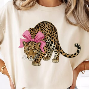 May include: Cream-colored t-shirt featuring a leopard illustration with a pink bow. The leopard has a spotted coat and is depicted in a crouching pose. The design is centered on the shirt, creating a playful and stylish look.