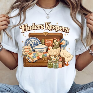 Könnte beinhalten: Weißes T-Shirt mit dem Text "Finders Keepers" in einer Retro-Schriftart. Die Grafik zeigt eine Holzkiste voller Vintage-Artikel, darunter eine Steppdecke, Geschirr, Kochbücher und eine Ente.