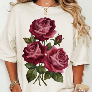 Może przedstawiać: Kremowy t-shirt z szczegółową ilustracją trzech czerwonych róż z zielonymi liśćmi i pąkami. Róże mają gradient czerwieni i różu. Koszulka ma luźny krój z krótkimi rękawami, odpowiednia do noszenia na co dzień.