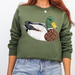 May include: An olive green sweatshirt featuring a detailed appliqu&eacute; of a duck. The duck has a green and black plaid head, a yellow beak, and wings with patterned fabric in shades of black, white, and brown. The sweatshirt is a casual style.