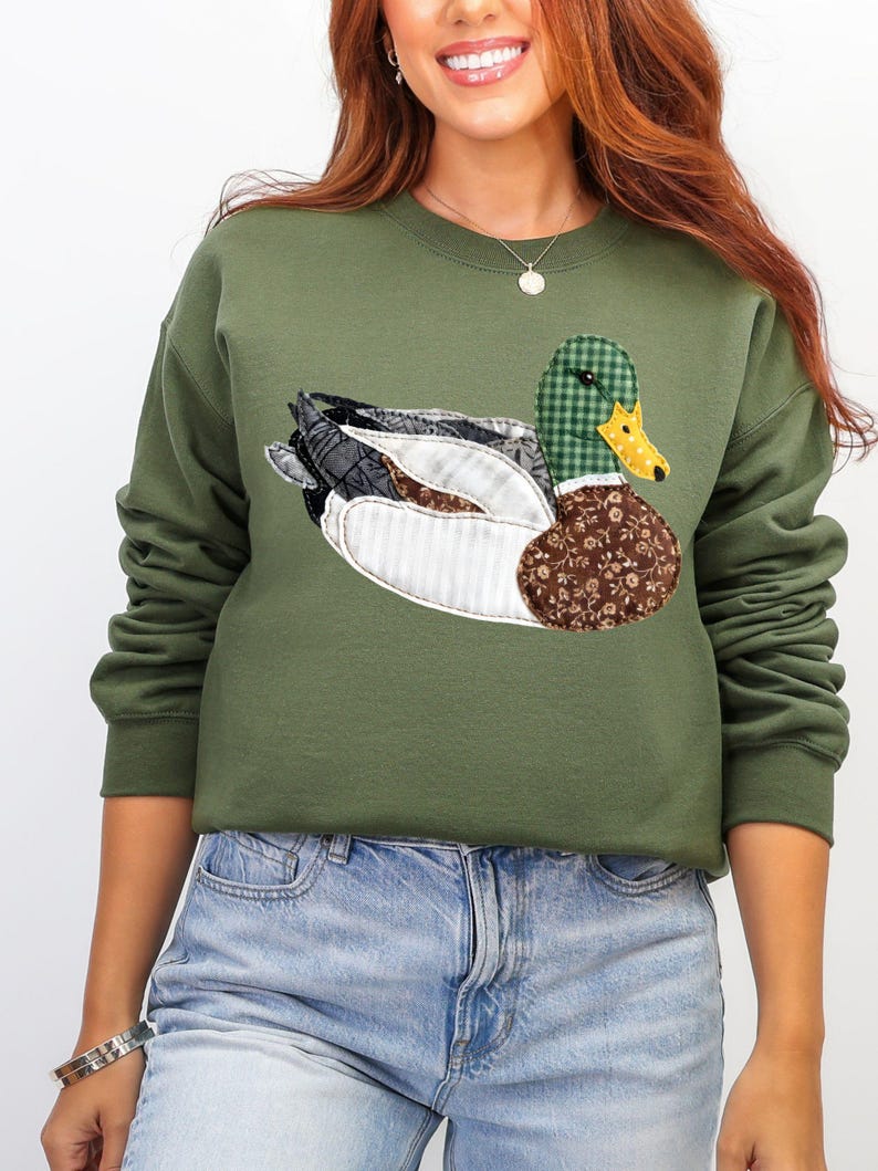 May include: An olive green sweatshirt featuring a detailed appliqu&eacute; of a duck. The duck has a green and black plaid head, a yellow beak, and wings with patterned fabric in shades of black, white, and brown. The sweatshirt is a casual style.