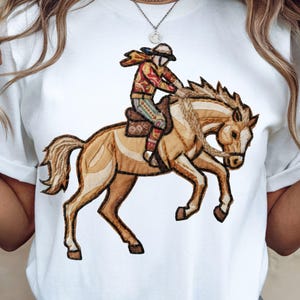 May include: White t-shirt featuring an embroidered design of a rider on a horse. The rider wears a red and yellow patterned jacket and hat. The horse is brown and tan with a braided mane and tail.
