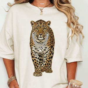 May include: Cream-colored t-shirt featuring a detailed illustration of a leopard wearing a pearl necklace and earrings. The leopard has a spotted coat and is centered on the shirt.