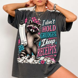 K&ouml;nnte beinhalten: Ein anthrazitfarbenes T-Shirt mit einer Grafik eines Waschb&auml;ren, der eine rosa Sonnenbrille tr&auml;gt und einen langen Kassenbon h&auml;lt. Der Text lautet: I don't HOLD GRUDGES. I keep RECEIPTS. Das Design enth&auml;lt rosa Blitze, blaue Karomuster und Herzen.