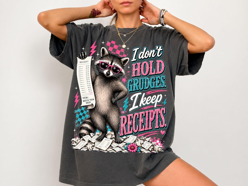 K&ouml;nnte beinhalten: Ein anthrazitfarbenes T-Shirt mit einer Grafik eines Waschb&auml;ren, der eine rosa Sonnenbrille tr&auml;gt und einen langen Kassenbon h&auml;lt. Der Text lautet: I don't HOLD GRUDGES. I keep RECEIPTS. Das Design enth&auml;lt rosa Blitze, blaue Karomuster und Herzen.