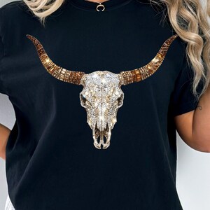 May include: A black short-sleeved t-shirt featuring a central graphic of a bull skull. The skull is decorated with clear, white, and amber-colored rhinestones that create a sparkling, multi-faceted appearance across the horns and cranium.