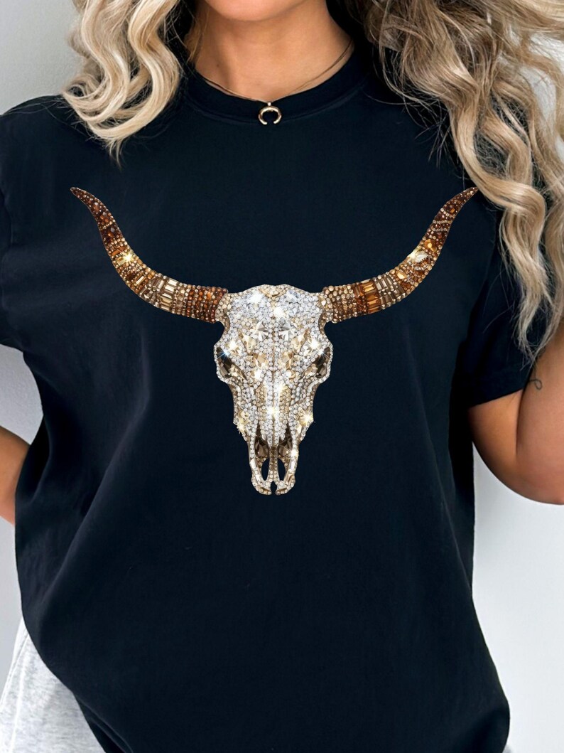 May include: A black short-sleeved t-shirt featuring a central graphic of a bull skull. The skull is decorated with clear, white, and amber-colored rhinestones that create a sparkling, multi-faceted appearance across the horns and cranium.