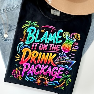 May include: Black t-shirt with a colorful graphic that reads "Blame it on the Drink Package." The design includes a cocktail, cruise ship, palm trees, and a sunset. The shirt is paired with blue jeans and a tan hat.
