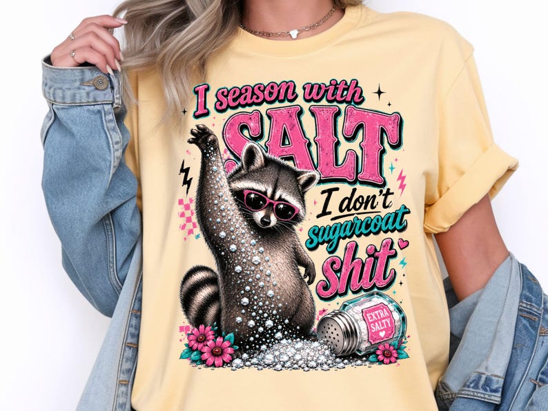 May include: A pale yellow t-shirt featuring a raccoon wearing sunglasses and the text "I season with SALT I don't sugarcoat Shit". The design includes salt, flowers, and a salt shaker.