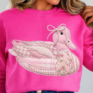 May include: A bright pink sweatshirt featuring a patchwork duck design. The duck is composed of various pink fabrics, including plaid, polka dots, and floral patterns. The duck has a bow on its head and is centered on the front of the shirt.