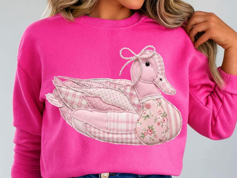 May include: A bright pink sweatshirt featuring a patchwork duck design. The duck is composed of various pink fabrics, including plaid, polka dots, and floral patterns. The duck has a bow on its head and is centered on the front of the shirt.