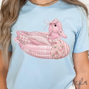 May include: Light blue t-shirt featuring a pink duck design. The duck is made of patchwork fabric with various patterns, including plaid, polka dots, and floral prints. A small bow adorns the duck's head, adding a touch of whimsy. The shirt is a comfortable, casual wear.