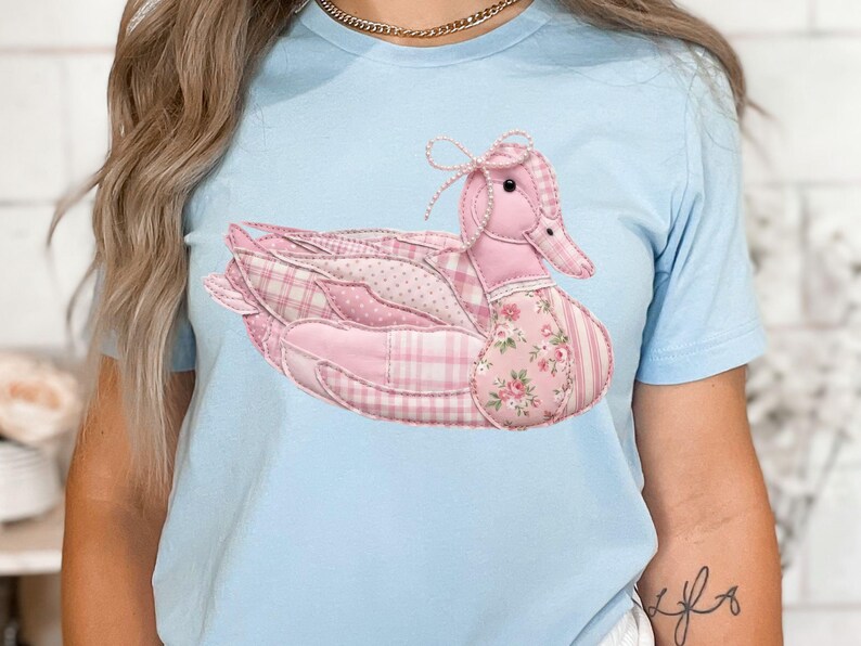 May include: Light blue t-shirt featuring a pink duck design. The duck is made of patchwork fabric with various patterns, including plaid, polka dots, and floral prints. A small bow adorns the duck's head, adding a touch of whimsy. The shirt is a comfortable, casual wear.