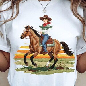 Peut inclure: T-shirt blanc avec un motif d'une personne montant un cheval brun. La personne porte un chapeau de cowboy, un foulard rouge et un jean bleu. Le fond présente un coucher de soleil avec de l'herbe verte.