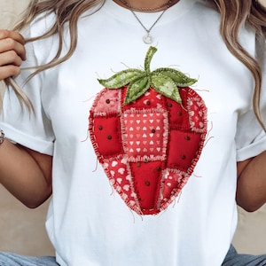 May include: White t-shirt featuring a large, patchwork-style strawberry design. The strawberry is primarily red with green leaves and a stem. The design incorporates various red fabric patches with heart patterns.