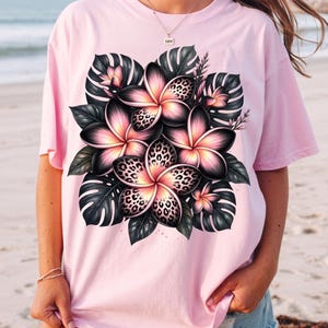 Può includere: Una maglietta rosa chiaro con un motivo floreale. Il design include fiori rosa con centri a stampa leopardata, circondati da foglie verde scuro. La maglietta è di colore rosa chiaro.