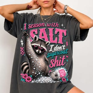 May include: Dark grey t-shirt featuring a raccoon in sunglasses, holding a salt shaker. Text says "I season with SALT, I don't sugarcoat shit." Pink and turquoise accents with pink flowers.