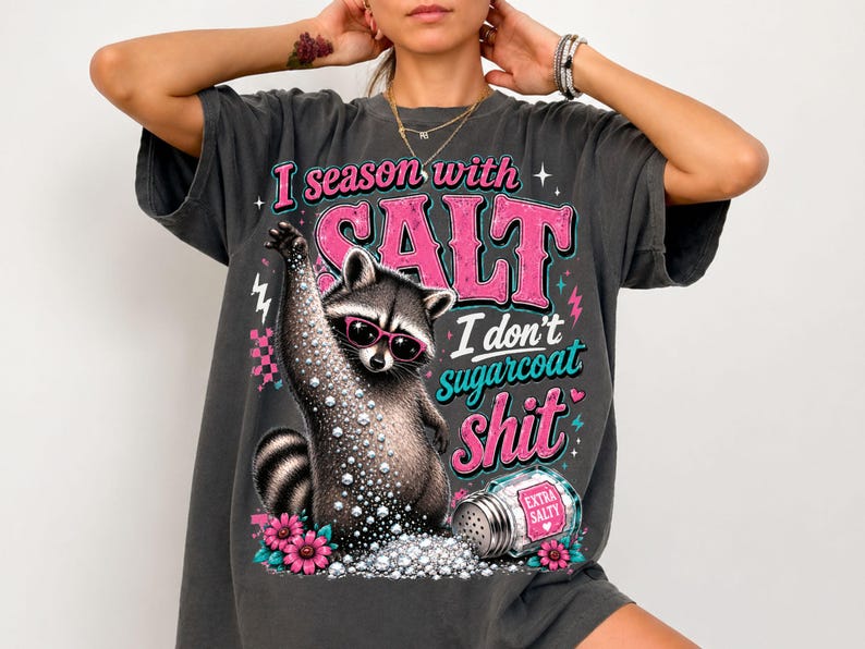 May include: Dark grey t-shirt featuring a raccoon in sunglasses, holding a salt shaker. Text says "I season with SALT, I don't sugarcoat shit." Pink and turquoise accents with pink flowers.
