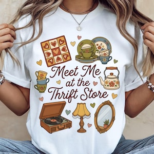 Op de afbeelding: Wit T-shirt met een grafisch ontwerp met vintage kringloopartikelen zoals een platenspeler, een theeset en een spiegel. De tekst "Meet Me at the Thrift Store" is ook in het ontwerp opgenomen.
