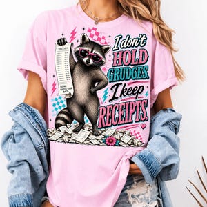 K&ouml;nnte beinhalten: Ein hellrosa T-Shirt mit einer Grafik eines Waschb&auml;ren, der eine rosa Sonnenbrille tr&auml;gt und einen langen Kassenbon h&auml;lt. Der Text lautet, I don't HOLD GRUDGES. I keep RECEIPTS. Das Design enth&auml;lt rosa Blitze, blaue Karomuster und einen Haufen Kassenbons.