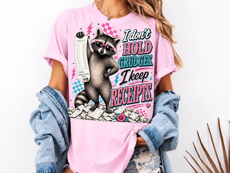 K&ouml;nnte beinhalten: Ein hellrosa T-Shirt mit einer Grafik eines Waschb&auml;ren, der eine rosa Sonnenbrille tr&auml;gt und einen langen Kassenbon h&auml;lt. Der Text lautet, I don't HOLD GRUDGES. I keep RECEIPTS. Das Design enth&auml;lt rosa Blitze, blaue Karomuster und einen Haufen Kassenbons.