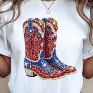 Puede incluir: Una camiseta blanca con un gráfico de un par de botas de vaquero ornamentadas. Las botas son rojas, blancas y azules con adornos de estrellas y joyas. Las botas tienen ribetes dorados y tacones plateados.