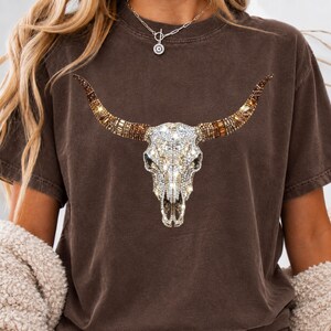 May include: A dark brown short-sleeve t-shirt featuring a large graphic of a bull skull. The skull is decorated with a sparkling, multi-faceted crystal and rhinestone design in shades of clear, gold, and bronze, covering the entire surface.