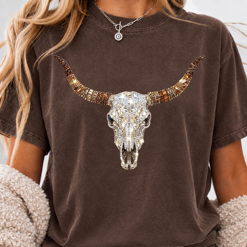 May include: A dark brown short-sleeve t-shirt featuring a large graphic of a bull skull. The skull is decorated with a sparkling, multi-faceted crystal and rhinestone design in shades of clear, gold, and bronze, covering the entire surface.