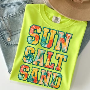 May include: A neon green t-shirt with the words "SUN SALT SAND" in a colorful floral design. The shirt is folded and placed on top of a pair of light-wash denim jeans and a tan hat.