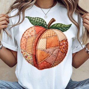 May include: White t-shirt featuring a patchwork peach design. The peach is composed of various fabric patterns in shades of orange, red, and yellow, with green leaves and a brown stem. The shirt has short sleeves and a round neckline.