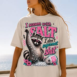 May include: A cream-coloured t-shirt with a raccoon wearing sunglasses and surrounded by salt. The text reads "I season with SALT I don't sugarcoat shit." The design includes pink and teal accents. The image is set against a blue sky.