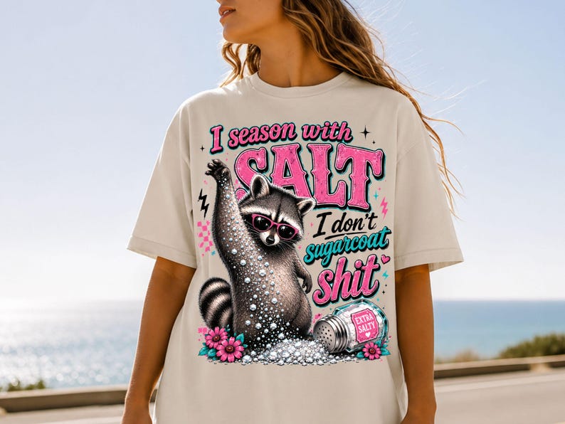 May include: A cream-coloured t-shirt with a raccoon wearing sunglasses and surrounded by salt. The text reads "I season with SALT I don't sugarcoat shit." The design includes pink and teal accents. The image is set against a blue sky.