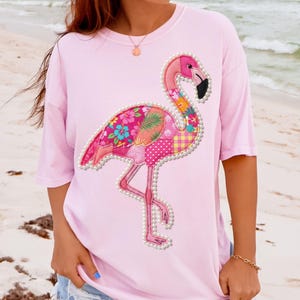 May include: A light pink t-shirt featuring a flamingo design. The flamingo is adorned with a patchwork of floral and polka dot patterns in shades of pink, orange, and yellow, outlined with faux pearls. The flamingo has a black beak.