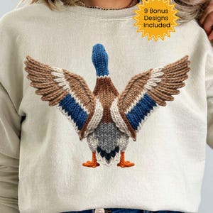 May include: A cream-colored sweatshirt featuring a detailed knitted duck design with outstretched wings. The duck has brown, blue, and white accents. The text "9 Bonus Designs Included" is in a yellow starburst.