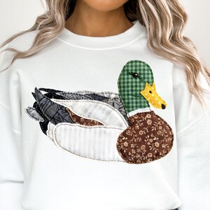 May include: White sweatshirt featuring a large, appliqued duck design. The duck has a green checkered head, a yellow beak, and a brown floral patterned body. The wings are a mix of patterned fabrics. A casual, nature-themed garment.