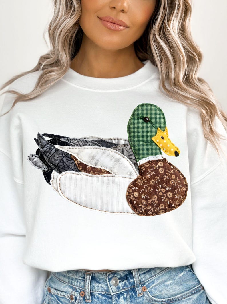 May include: White sweatshirt featuring a large, appliqued duck design. The duck has a green checkered head, a yellow beak, and a brown floral patterned body. The wings are a mix of patterned fabrics. A casual, nature-themed garment.