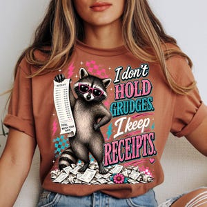K&ouml;nnte beinhalten: Ein rostfarbenes T-Shirt mit einer Grafik eines Waschb&auml;ren, der eine rosa Sonnenbrille tr&auml;gt und einen langen Kassenbon h&auml;lt. Der Text lautet: I don't hold grudges. I keep receipts. Total: Watch your back. Mit rosa und blauen Akzenten.