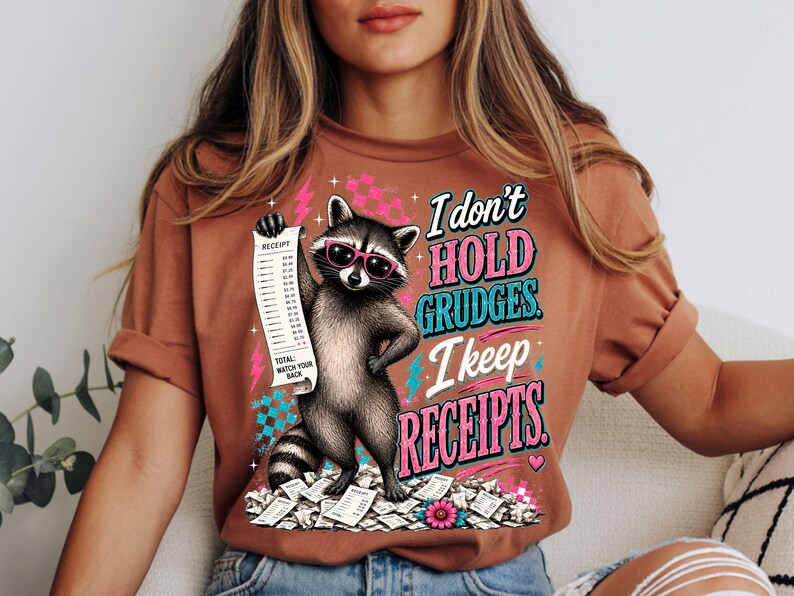 K&ouml;nnte beinhalten: Ein rostfarbenes T-Shirt mit einer Grafik eines Waschb&auml;ren, der eine rosa Sonnenbrille tr&auml;gt und einen langen Kassenbon h&auml;lt. Der Text lautet: I don't hold grudges. I keep receipts. Total: Watch your back. Mit rosa und blauen Akzenten.