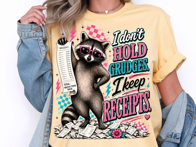 K&ouml;nnte beinhalten: Ein hellgelbes T-Shirt mit einer Grafik eines Waschb&auml;ren, der eine rosa Sonnenbrille tr&auml;gt und einen langen Kassenbon h&auml;lt. Der Text lautet, I don't hold grudges. I keep receipts. Das Design enth&auml;lt rosa und blaue Blitze sowie verstreute Kassenbons.