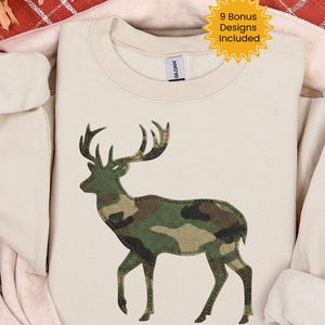 Pode incluir: Um moletom creme com um design de veado em padrão de camuflagem. O veado é retratado em uma pose em pé com grandes chifres. O texto "9 Bonus Designs Included" está em uma estrela amarela.