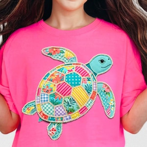 May include: A bright pink t-shirt featuring a colorful turtle applique. The turtle has a light blue head and limbs, and a patchwork shell with floral and geometric patterns. The applique is outlined with small white beads.